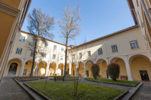 Dziedziniec uniwersytecki. Zdjęcie wewnętrznego, prostokątnego dziedzińca otoczonego arkadami i żółtymi murami budynku, z ławkami i kilkoma drzewkami, oświetlonego słonecznym światłem.