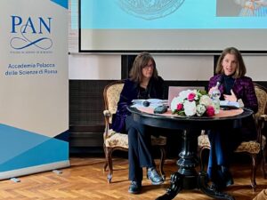 Panel dyskusyjny. Dwie kobiety siedzą przy okrągłym stoliku w eleganckich, tapicerowanych fotelach – jedna z nich trzyma mikrofon i gestykuluje. Z lewej stoi stojak z banerem PAN (Polish Academy of Sciences), w tle fragment ekranu projekcyjnego.