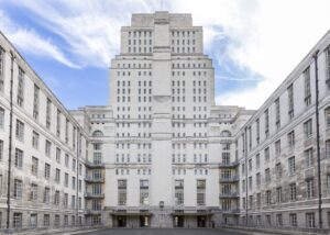 Gmach Senate House, University of London. Symetryczny, wysokiego art déco budynek z jasnego kamienia, z centralną wieżą i skrzydłami po bokach, na tle niebieskiego, lekko zachmurzonego nieba.