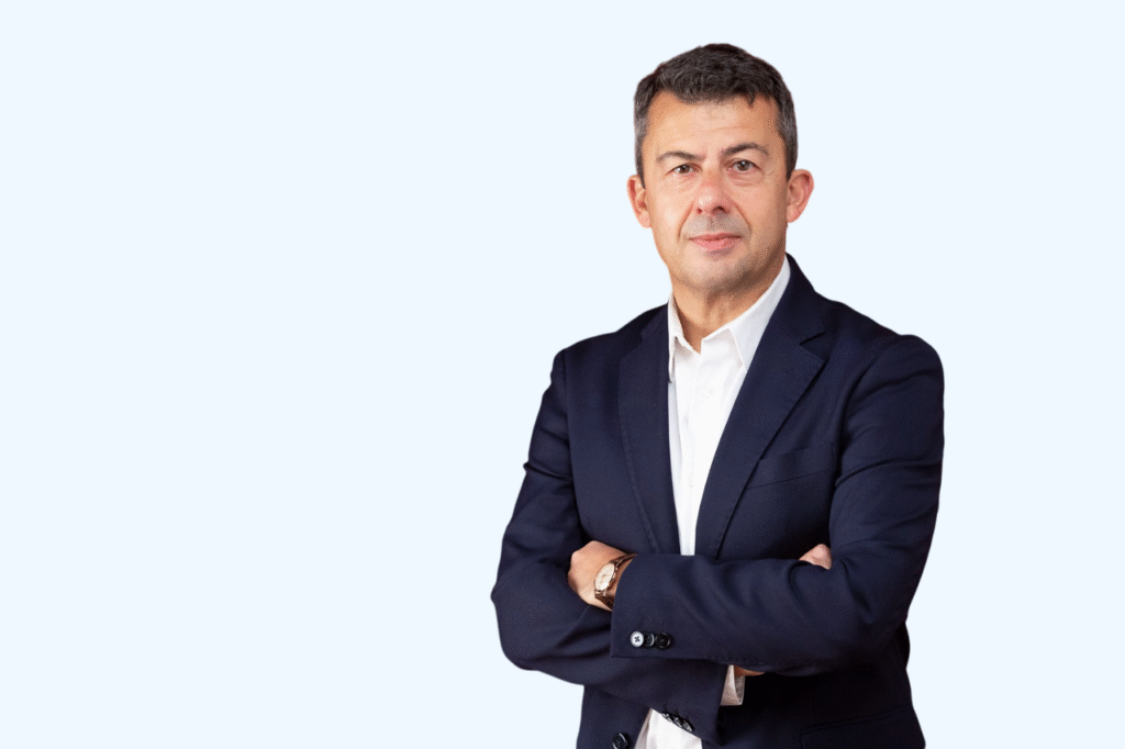Mężczyzna w ciemnym marynarce i białej koszuli, ze skrzyżowanymi rękami, na jednolitym, jasnoniebieskim tle, patrzy w obiektyw.